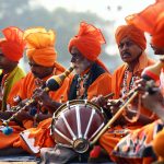 A group of Indian men in vibrant orange traditional attire performing music during a cultural festival outdoors.